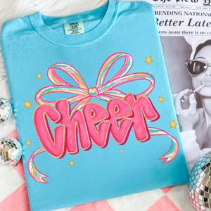 May include: A light blue t-shirt with the word "Cheer" in pink, outlined in gold, and a colorful bow design. The shirt also features small yellow star accents. The shirt is made of a soft material.