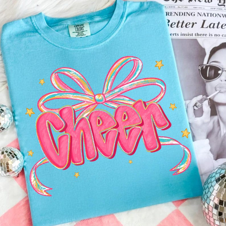 May include: A light blue t-shirt with the word "Cheer" in pink, outlined in gold, and a colorful bow design. The shirt also features small yellow star accents. The shirt is made of a soft material.