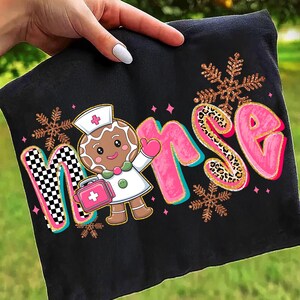 May include: Black fabric with the word "NURSE" in pink, gold, and checkered letters, featuring a gingerbread nurse with a pink medical bag and snowflakes. The design is held by a hand.
