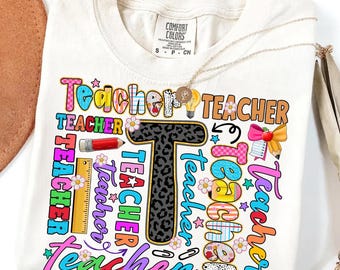 以下が含まれることがあります： クリーム色のTシャツに、カラフルで気まぐれなデザインで「Teacher」の文字がプリントされています。「T」の文字はヒョウ柄です。金のネックレスがシャツにかけられています。シャツはComfort Colorsブランドです。