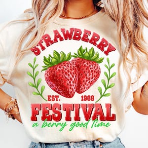 May include: A white t-shirt with a red graphic that says "Strawberry Festival Est. 1968 a berry good time". The graphic also features two red strawberries with green leaves.