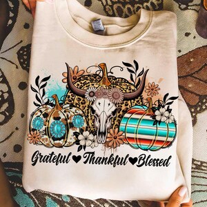 Puede incluir: Sudadera crema con un dise&ntilde;o de calabazas, una calavera de vaca y flores. Las calabazas tienen patrones turquesa, de leopardo y a rayas. El texto "Grateful Thankful Blessed" est&aacute; impreso debajo.