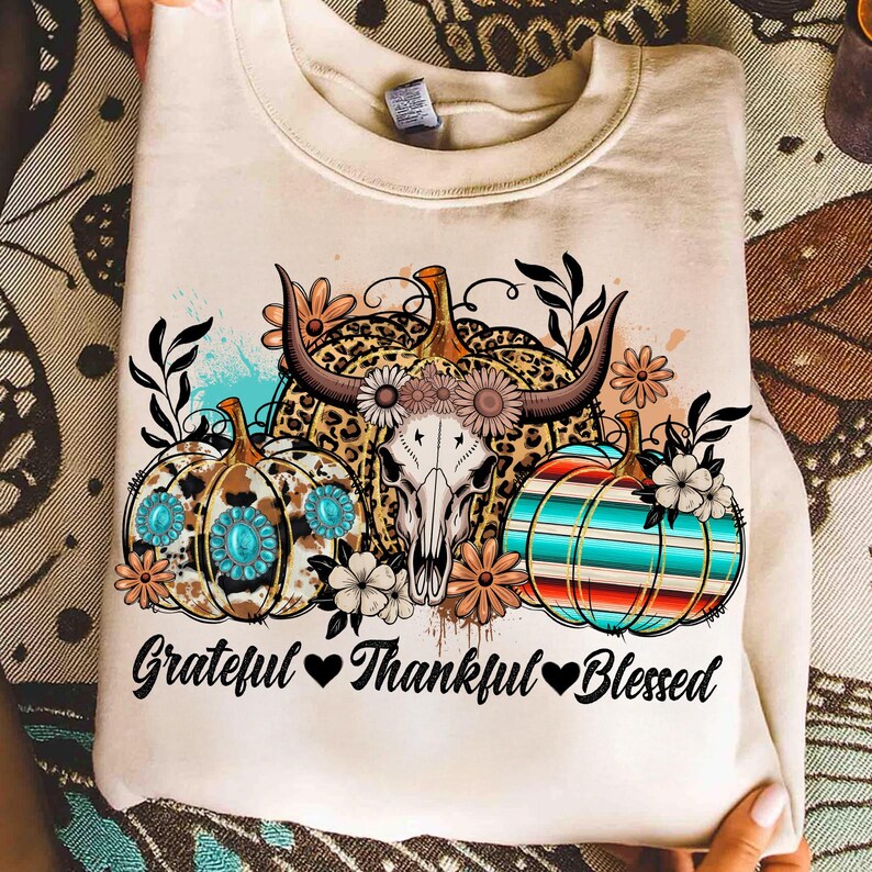 Puede incluir: Sudadera crema con un dise&ntilde;o de calabazas, una calavera de vaca y flores. Las calabazas tienen patrones turquesa, de leopardo y a rayas. El texto "Grateful Thankful Blessed" est&aacute; impreso debajo.