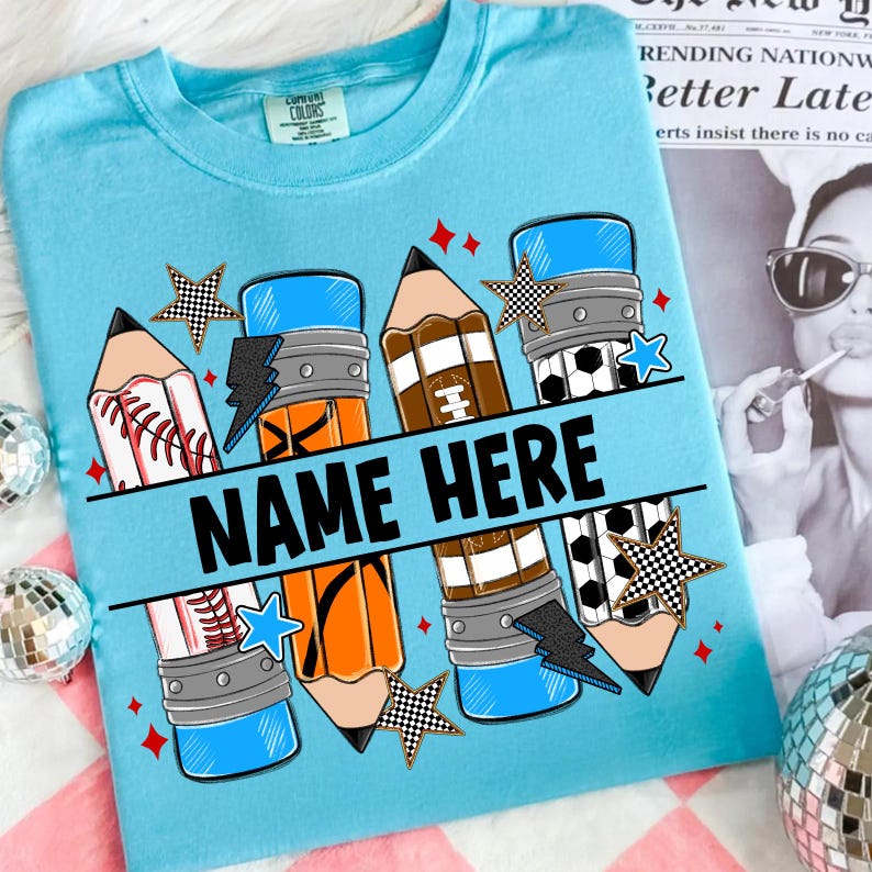 Pode incluir: Camiseta azul clara com um design gr&aacute;fico com l&aacute;pis com desenhos de bolas esportivas, raios e estrelas. O texto "NAME HERE" &eacute; exibido em letras pretas em negrito no centro da camiseta.