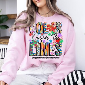 May include: Pink sweatshirt with a colorful graphic design that says "Color Outside The Lines #AutismAwareness".