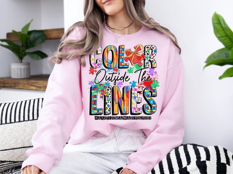 May include: Pink sweatshirt with a colorful graphic design that says "Color Outside The Lines #AutismAwareness".