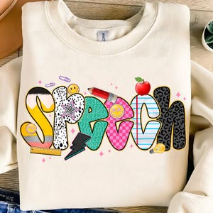 Peut inclure: Sweat-shirt crème avec le mot "SPEECH" en lettres colorées et ludiques. Les lettres présentent divers motifs, dont des rayures, un imprimé léopard et un crayon. Une pomme rouge et une ampoule sont également incluses dans le design.