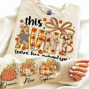 May include: Cream-colored sweatshirt with the text "this AUNT loves her pumpkins" in a fall-themed design. The word "AUNT" is decorated with plaid, floral, and star patterns. The sleeve features three pumpkin illustrations with names.