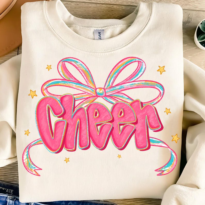 May include: Cream-colored sweatshirt with the word "Cheer" in large, bubble-style pink letters, accented with a rainbow-colored bow and ribbons. Small yellow stars are scattered around the design. The sweatshirt is made of a soft material.