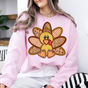 May include: A pink sweatshirt featuring a patchwork turkey design. The turkey is made from various fabrics, including plaid, leopard print, and leaves. The turkey has a yellow body, brown eyes, and a red beak.