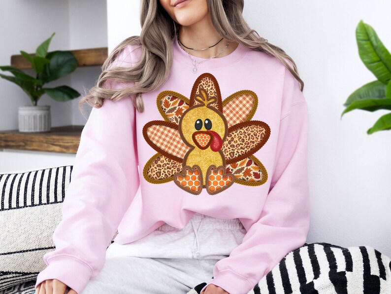 May include: A pink sweatshirt featuring a patchwork turkey design. The turkey is made from various fabrics, including plaid, leopard print, and leaves. The turkey has a yellow body, brown eyes, and a red beak.