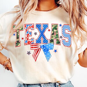 May include: A cream colored t-shirt with the word "TEXAS" printed in blue, red, and green letters. The letters are made up of different patterns, including camouflage, stars, and checkered designs. A red, white, and blue bow is centered below the word "TEXAS".