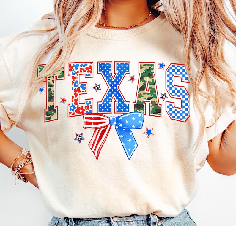 May include: A cream colored t-shirt with the word "TEXAS" printed in blue, red, and green letters. The letters are made up of different patterns, including camouflage, stars, and checkered designs. A red, white, and blue bow is centered below the word "TEXAS".