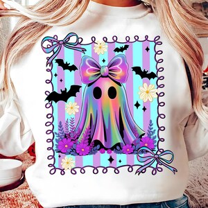 May include: White sweatshirt featuring a colorful ghost design with a large bow, bats, flowers, and a striped background. The design is framed with a decorative border and bows. A festive and whimsical Halloween-themed apparel.