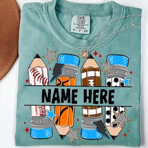 Pode incluir: Uma camiseta Comfort Colors verde-azulado com um design de l&aacute;pis com bolas esportivas e um raio. O texto "NAME HERE" &eacute; impresso em negrito preto. Um colar de prata com um pingente tamb&eacute;m &eacute; vis&iacute;vel.