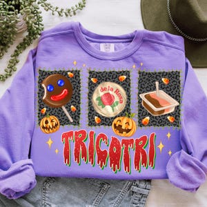 K&ouml;nnte beinhalten: Ein lila Sweatshirt mit einem Halloween-Motiv. Das Design umfasst drei Quadrate mit einem Lutscher, einer Rose und einem Dessert. Das Wort "TRICOTRI" ist in Rot und Gr&uuml;n geschrieben. Maisbonbons und K&uuml;rbisse sind ebenfalls abgebildet.