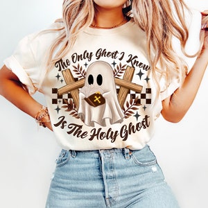 Op de afbeelding: Een wit T-shirt met een afbeelding van een geest die een boek met een kruis erop vasthoudt. De tekst op het shirt luidt "The Only Ghost I Know Is The Holy Ghost".