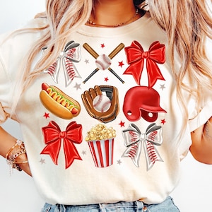 May include: A white t-shirt with a baseball theme. The design includes a baseball, bat, glove, helmet, hot dog, popcorn, and red bows.