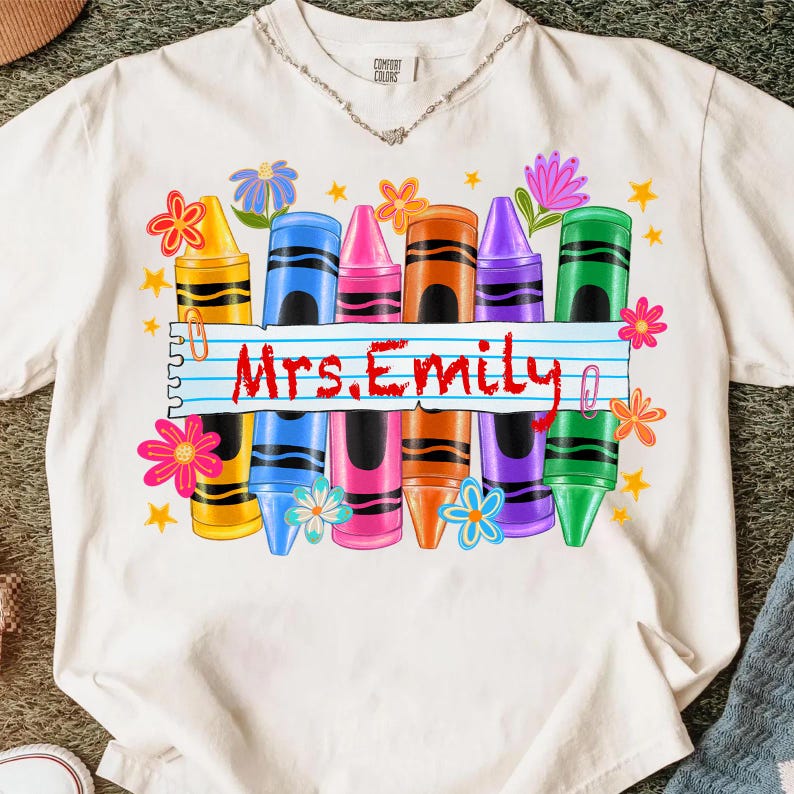 May include: White t-shirt featuring a colorful crayon design with the text "Mrs. Emily". The design includes yellow, blue, pink, orange, purple, and green crayons, along with floral and star accents. The shirt is a Comfort Colors brand.
