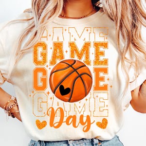 Peut inclure: T-shirt blanc avec un motif de ballon de basket orange et noir et le texte "Game Day" en lettres orange paillet&eacute;es.