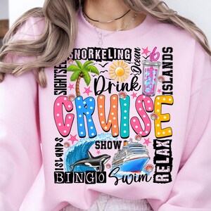 May include: Pink sweatshirt with a colorful graphic design that says "Drink Cruise" and includes images of a palm tree, a cruise ship, a whale, and a cocktail. The graphic also includes the words "Bingo", "Swim", "Show", "Relax", "Snorkeling", "Ocean", "Islands", and "Sights"