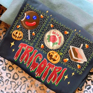K&ouml;nnte beinhalten: Dunkelblaues T-Shirt mit einem Halloween-Motiv. Das Shirt zeigt einen Lebkuchenmann-Lutscher, K&uuml;rbisse, Zuckermais und das Wort "TRICKORTREAT" in roter und gr&uuml;ner Schrift. Das Design ist von einem Leopardenmuster umgeben.