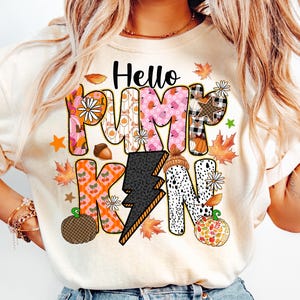 May include: Cream-colored t-shirt with the words "Hello Pumpkin" in a colorful, patterned design. The letters are filled with various autumn-themed patterns, including pumpkins, leaves, and plaid. Decorative elements include pumpkins, acorns, and stars.