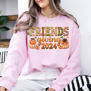 May include: Pink sweatshirt with the text "Friendsgiving 2024" in a plaid and leopard print font. The text is surrounded by small hearts and two illustrations of pumpkins.