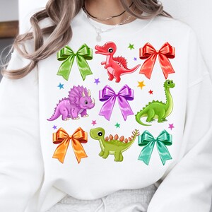 May include: White sweatshirt with a colorful dinosaur and bow design. The dinosaurs are green, red, purple, and green. The bows are pink, orange, green, and blue.