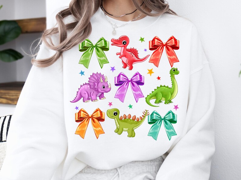 May include: White sweatshirt with a colorful dinosaur and bow design. The dinosaurs are green, red, purple, and green. The bows are pink, orange, green, and blue.