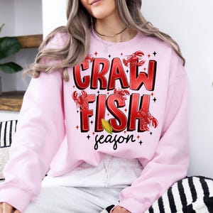 May include: Pink sweatshirt with a graphic design that says "Crawfish season" with red crawfish illustrations and a yellow corn cob.