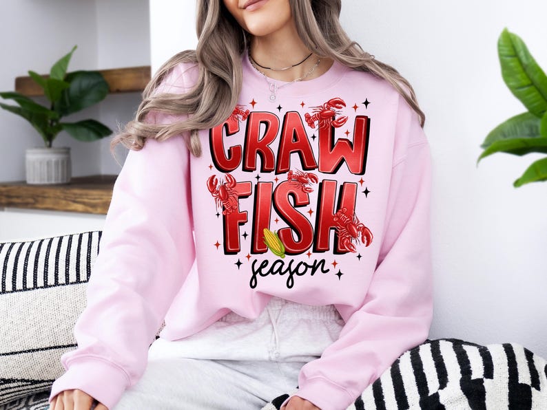 May include: Pink sweatshirt with a graphic design that says "Crawfish season" with red crawfish illustrations and a yellow corn cob.