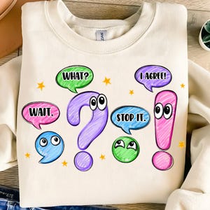 May include: Cream-colored sweatshirt with a whimsical design featuring a large purple question mark and pink exclamation point, both with cartoon eyes. Speech bubbles say "WAIT.", "WHAT?", "STOP IT.", and "I AGREE!"
