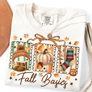 May include: Cream-colored t-shirt with a graphic design featuring fall-themed illustrations: a hat, boots, pumpkin, coffee, sock, and candle. The text "Fall Basics" is printed in a script font. A gold necklace is draped across the shirt.