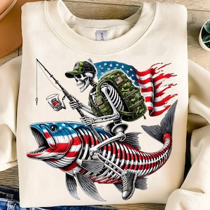 May include: Cream-colored sweatshirt featuring a skeleton angler riding a fish with an American flag design. The skeleton wears a camouflage backpack and hat, holding a fishing rod. The fish is red, white, and blue.
