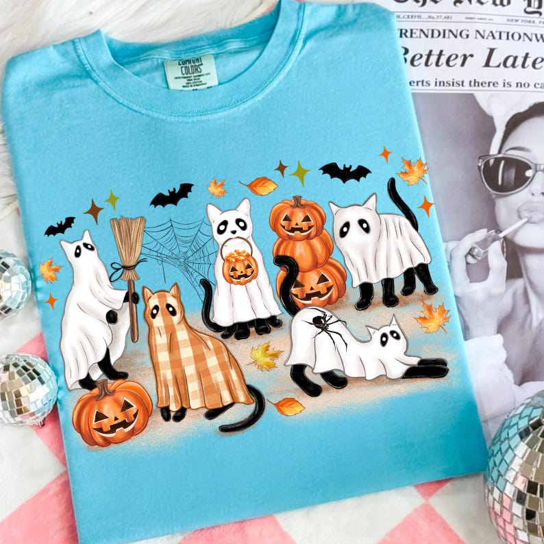 May include: A light blue Comfort Colors t-shirt with a Halloween design. The design features cartoon cats in ghost costumes, pumpkins, and a plaid cat, alongside bats, spiderwebs, and autumn leaves. The t-shirt has a crew neck.