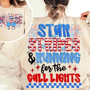 May include: A beige crewneck sweatshirt featuring a graphic print with the text STAR STRIPES & RUNNING for the CALL LIGHTS in red, white, and blue. The design includes fireworks, stars, and a checkered pattern, with a nurse-themed inset graphic.
