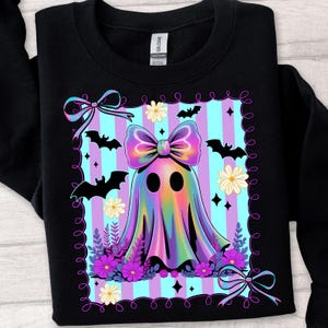 May include: Black sweatshirt featuring a colorful ghost design with a large bow, flowers, and bats. The ghost has a rainbow-colored body and is set against a striped background. The design includes decorative bows and a floral border, perfect for Halloween.