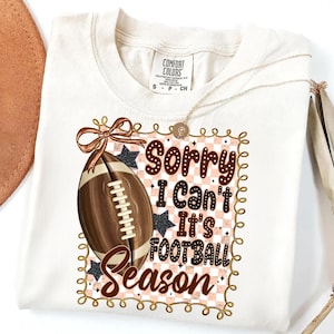 May include: Cream-colored t-shirt with a football graphic and the text "Sorry I Can't It's Football Season." The design includes a football, stars, and a decorative border. A gold necklace is draped across the shirt.