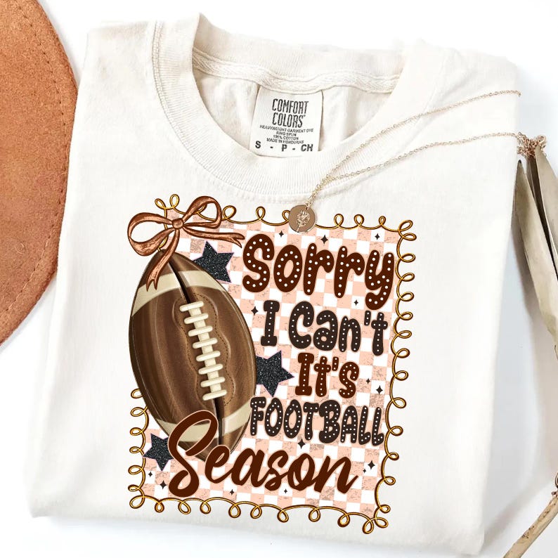 May include: Cream-colored t-shirt with a football graphic and the text "Sorry I Can't It's Football Season." The design includes a football, stars, and a decorative border. A gold necklace is draped across the shirt.