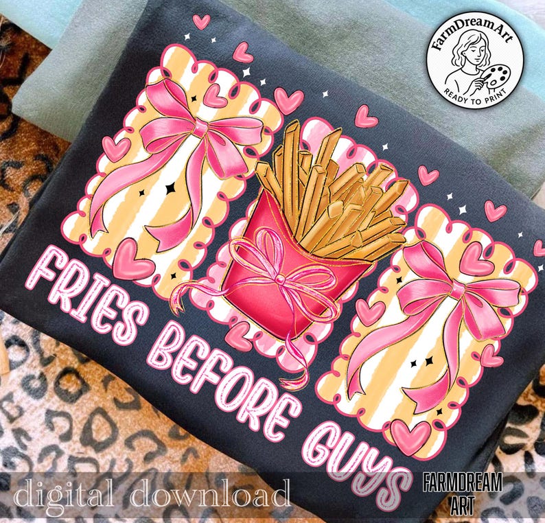 Op de afbeelding: Zwart t-shirt met een grafisch ontwerp. Het heeft een rode bak friet, roze strikken en de tekst "FRIES BEFORE GUYS". Het ontwerp bevat roze harten en een decoratieve rand met een gestreepte achtergrond.