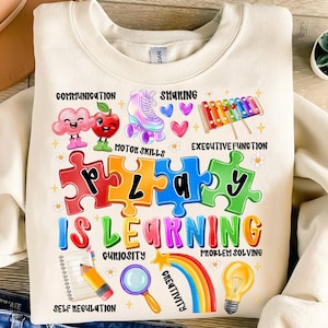 Puede incluir: Sudadera de color crema con un diseño gráfico colorido. El diseño incluye piezas de rompecabezas que deletrean "PLAY", las palabras "IS LEARNING" e ilustraciones de un patín, una manzana y un arcoíris. El texto adicional incluye "COMMUNICATION" y "CREATIVITY".