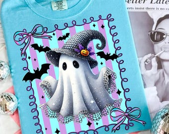 May include: Light blue t-shirt featuring a Halloween ghost wearing a witch hat with a pumpkin and bats. The ghost is surrounded by a purple and blue striped border with bows. The shirt has a crew neck and short sleeves.