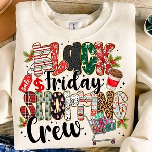May include: Cream-colored sweatshirt with a colorful graphic that reads "Black Friday Shopping Crew." The text incorporates various patterns, including stripes, plaid, and animal print, along with a shopping cart and coffee cup.