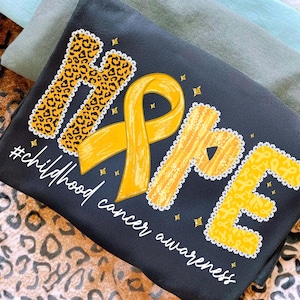 May include: Black t-shirt with the word "HOPE" in yellow and leopard print, with a yellow ribbon and the text "#childhood cancer awareness". The shirt is folded on top of other shirts and a leopard print fabric.