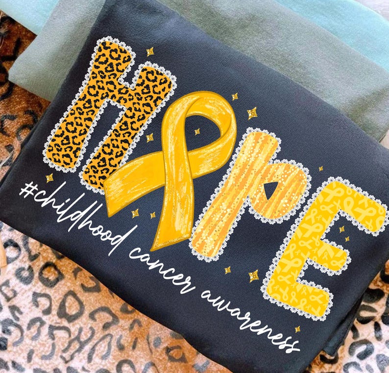 May include: Black t-shirt with the word "HOPE" in yellow and leopard print, with a yellow ribbon and the text "#childhood cancer awareness". The shirt is folded on top of other shirts and a leopard print fabric.