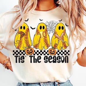 Puede incluir: Sudadera color crema con tres figuras de fantasmas amarillos con costuras de b&eacute;isbol, sosteniendo una pelota, un bate y un guante. Murci&eacute;lagos negros y una telara&ntilde;a est&aacute;n encima de los fantasmas. El texto "Tis' The Season" est&aacute; debajo, con calabazas.