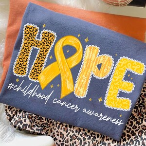 May include: A navy blue t-shirt with the word "HOPE" in yellow, leopard print, and ribbon designs. The shirt includes a yellow ribbon and the text "#childhood cancer awareness" in white. The shirt is folded on top of an orange shirt and leopard print fabric.