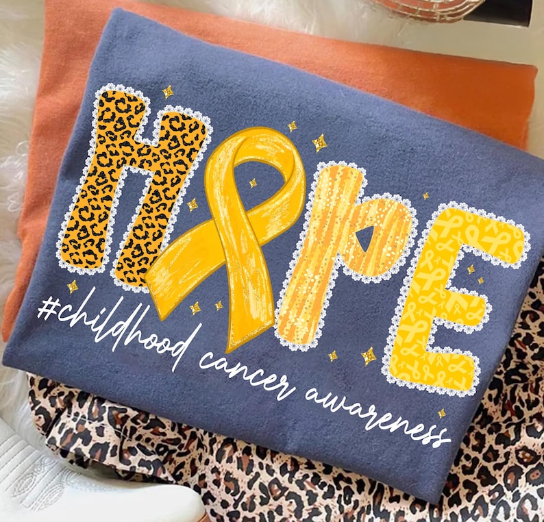 May include: A navy blue t-shirt with the word "HOPE" in yellow, leopard print, and ribbon designs. The shirt includes a yellow ribbon and the text "#childhood cancer awareness" in white. The shirt is folded on top of an orange shirt and leopard print fabric.