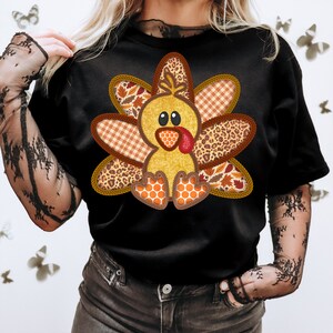 May include: A black t-shirt featuring a cute cartoon turkey design. The turkey is made from various patterned fabrics, including plaid, leopard print, and leaves. The turkey is wearing a red wattle and has a yellow body.
